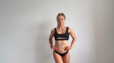 Siyah mayo giymiş genç, güzel, zinde ve doğal sarışın bir kız beyaz arka planda poz veriyor. Spor, fitness, cilt bakımı, selülit aldırma, diyet ve kilo kaybı.