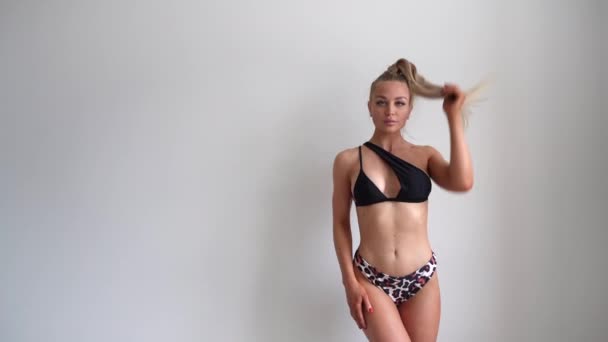 Una giovane, bella, in forma e naturale ragazza bionda in un costume da bagno leopardo nero posa su uno sfondo bianco con una coda di cavallo sulla testa. Sport, fitness, cura della pelle, rimozione della cellulite, dieta e perdita di peso — Video Stock