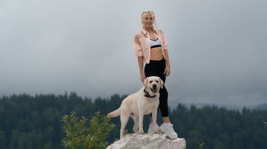 Sarışın bir kız, Labrador köpeğiyle birlikte kayanın üzerinde duruyor. Mutlu köpek. Ormanın arka planında yağmurlu bir gökyüzü