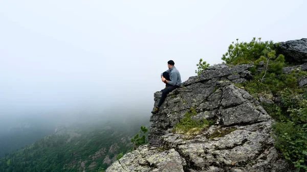 Мужчина сидит у обрыва на скале в тумане в горах летом — стоковое фото