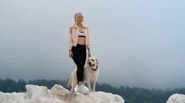 Sarışın bir kız, yavru beyaz bir Labrador köpeğiyle bir kayanın üzerinde duruyor. Ormanın arka planında yağmurlu bir gökyüzü