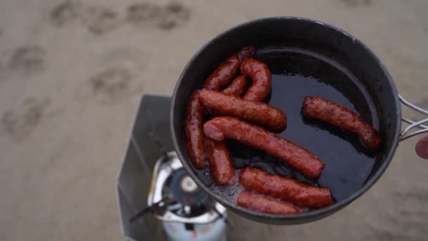 Hunting sausages in a frying pan are fried on a gas burner .against the backdrop of the beach and mountains holding a hand in the summer camping camp — Stock Video