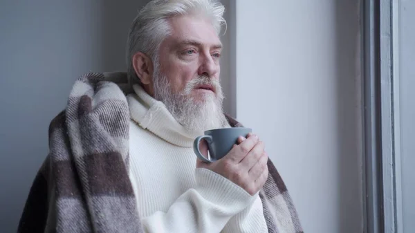 Starszy mężczyzna spogląda przez okno pije kawę w domu pokrytą ciepłym kocem — Zdjęcie stockowe