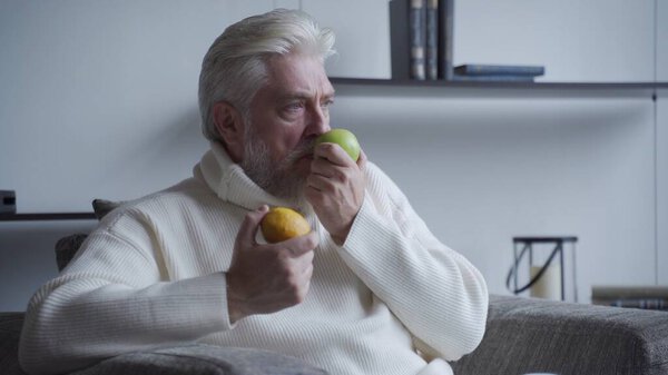 Elderly man with a gray beard sniffs lemon and Apple and does not smell