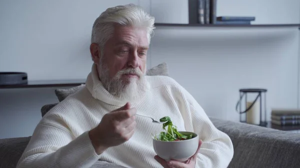 Starszy człowiek czuje się szczęśliwy jedząc dietetyczne jedzenie świeże sałatki na kanapie — Zdjęcie stockowe