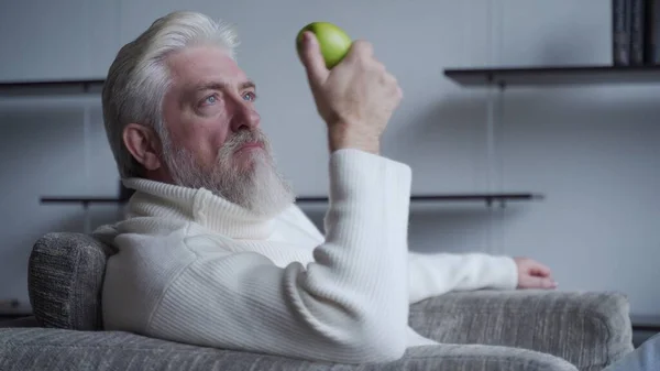Starszy mężczyzna z brodą siedzi sam na krześle trzymając jabłko w dłoni, myśląc — Zdjęcie stockowe