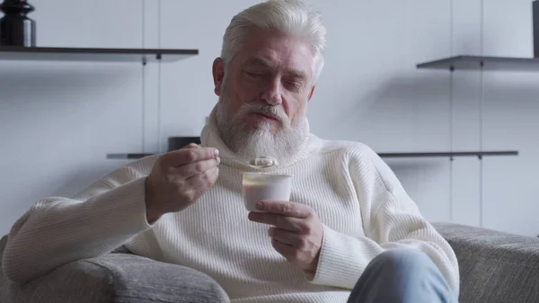 Красивый пожилой мужчина с седой бородой ест йогурт, держа ложку у рта. — стоковое фото
