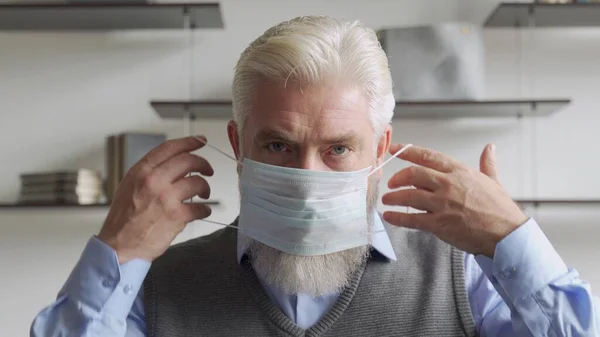 肖像画を閉じます高齢者の男性は自分自身を守るために彼の顔に医療マスクを置きますコロナウイルスパンデミックから. — ストック写真