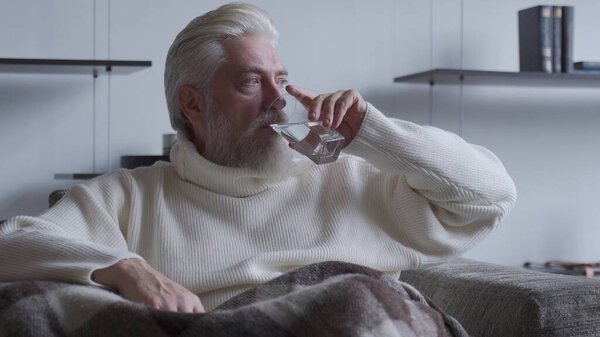 Elderly man with a gray beard is sitting in a chair and drinking a glass of water