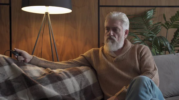 En äldre man med grått skägg sitter på soffan och tänder och släcker lampan. En grubblande äldre man är deprimerad — Stockfoto