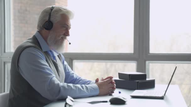 Пожилой бизнесмен в наушниках проводит видеоконференцию с деловым партнером, работающим в офисе — стоковое видео