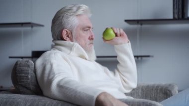 Sakallı yaşlı bir adam sandalyesinde tek başına oturuyor ve elinde bir elma olduğunu düşünüyor.