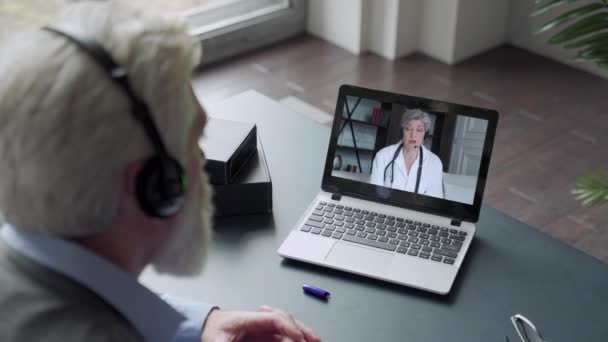 Um homem idoso com barba se comunica via computador com um médico via link de vídeo. Cuidados médicos em quarentena. Consulta remota com um terapeuta — Vídeo de Stock