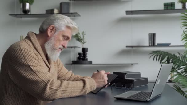 Um homem idoso com uma barba cinza desenha com uma caneta em um tablet em casa na sala de estar — Vídeo de Stock