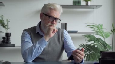 Gri sakallı yaşlı bir adam ofiste akıllı telefon kullanıyor.