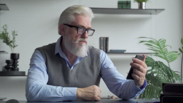 Un homme âgé aux cheveux gris avec une barbe grise est assis à l'intérieur, utilise un smartphone, vérifie les e-mails, lit des sms sur une vente incroyable, se sent heureux. Grandes réductions, grande offre, bonnes nouvelles, gagnants de loterie — Video