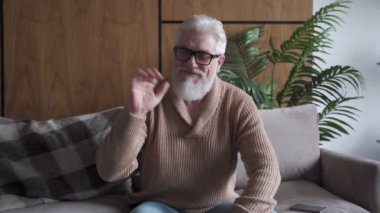Gri sakallı, mutlu yaşlı adam web kamerasıyla konuşuyor, gülümsüyor, yetişkin blogger kaydı yapıyor, kameraya bakıyor, evdeki yaşlı insanlarla çevrimiçi iletişim kuruyor.