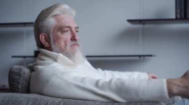 Sakallı yaşlı bir adam sandalyesinde tek başına oturuyor ve elinde bir elma olduğunu düşünüyor.