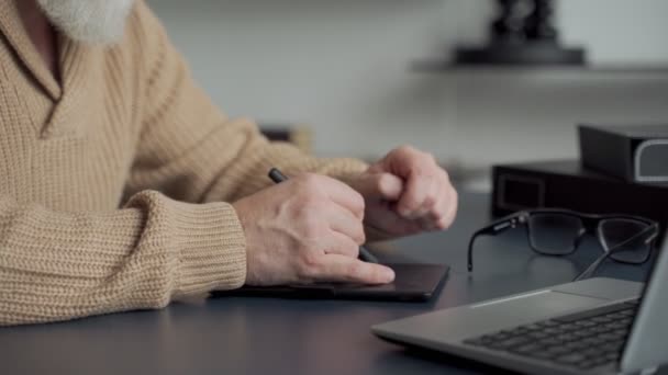 Um homem idoso com uma barba cinza desenha com uma caneta em um tablet em casa na sala de estar — Vídeo de Stock