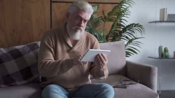 Notícias de leitura. Homem sênior com barba cinza usando tablet digital sentado no sofá em casa — Vídeo de Stock