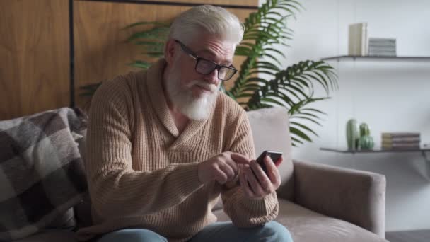 Feliz anciano sentado en el sofá mira con entusiasmo la pantalla del teléfono inteligente, celebrando la victoria del éxito o leyendo buenas noticias. — Vídeo de stock