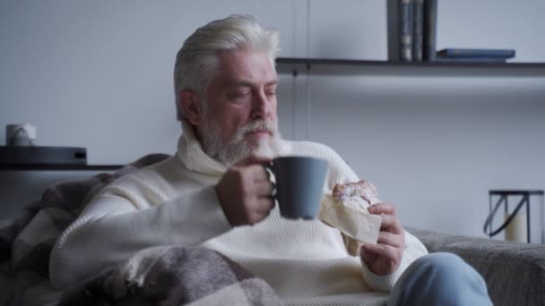 Anciano con barba gris está sentado en una silla bebiendo café y comiendo un croissant — Vídeos de Stock