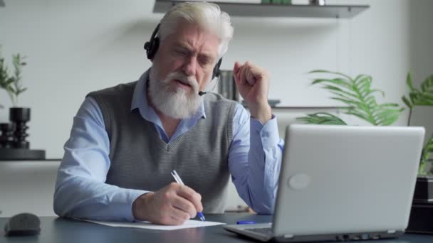 Lächelnder älterer Mann mit grauem Bart, der Kopfhörer trägt, einen Videoanruf auf einem Laptop hat, glücklicher älterer Mann mit Kopfhörern, der im Büro sitzt und mit moderner Technologie und drahtlosem Internet spricht — Stockvideo