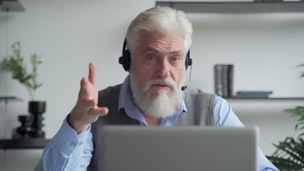 Lächelnder älterer Mann mit grauem Bart, der Kopfhörer trägt, einen Videoanruf auf einem Laptop hat, glücklicher älterer Mann mit Kopfhörern, der im Büro sitzt und mit moderner Technologie und drahtlosem Internet spricht — Stockvideo