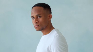 Siyah Afro-Amerikan bir adamın ciddi bir ifadeyle ışık stüdyosunun duvarına karşı poz verirken doğrudan kameraya baktığını düşünüyor.. 