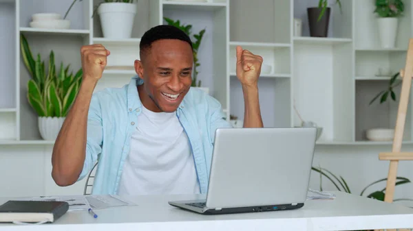 African-American feels happy received great news by internet looks at pc screen raised hands scream with joy celebrating on-line lottery win