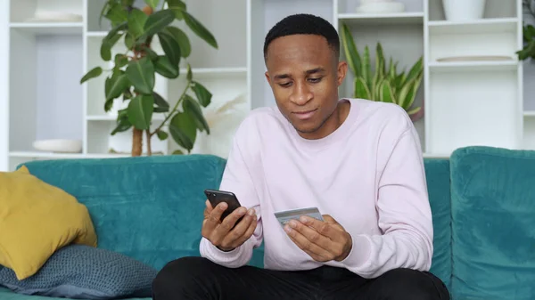 Pago en línea. Hombre afroamericano con tarjeta de crédito y el uso de smartphone para ir de compras en Internet en casa — Foto de Stock