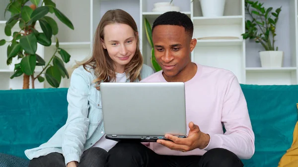 Cheerful interracial couple web surfing on laptop together, sitting on sofa at home