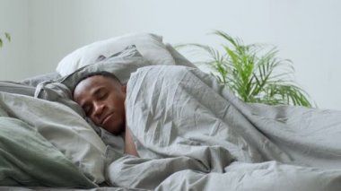 Genç siyahi adam yatak odasında uyanıyor. Mutlu Afrikalı adam sabahları yatakta geriniyor. Genç yetişkin uyandıktan sonra gülümsüyor..