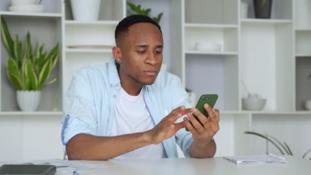 Genç melez ırk adamı masada oturuyor, akıllı telefondan SMS okuyor. Mobil uygulamalardaki e-postaları kontrol eden, ortaklarla iletişim kuran bin yıllık iş adamı. — Stok video