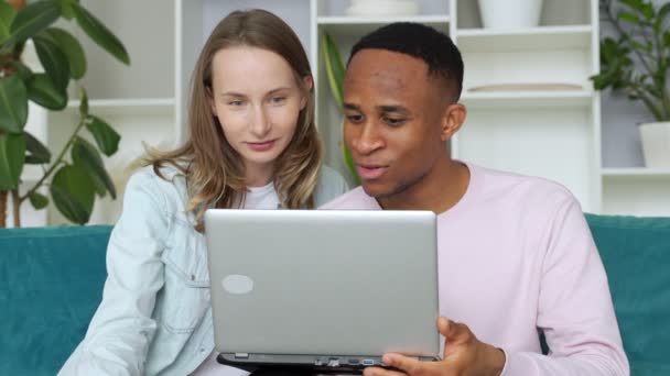 Pasangan ceria interrasial internet berselancar di laptop bersama-sama, duduk di sofa di rumah — Stok Video