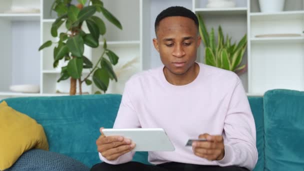 El hombre negro sonríe felizmente, hace compras en línea usando un dispositivo digital, y hace pagos bancarios en línea usando una tarjeta de débito o crédito — Vídeos de Stock