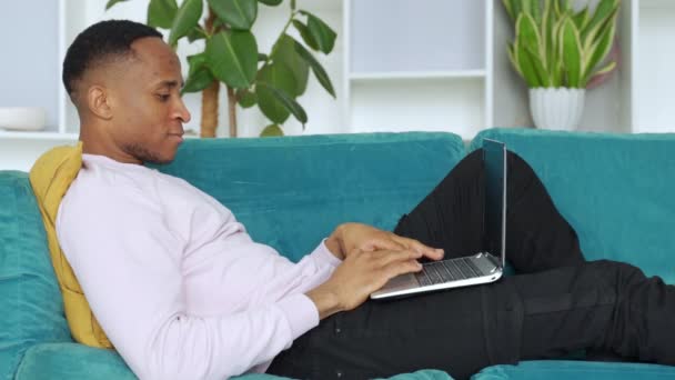 Siyah adam oturma odasında kanepede oturuyor ve internette arama yapmak, internette sörf yapmak ya da mesajlaşma yapmak için dizüstü bilgisayar kullanıyor.. — Stok video