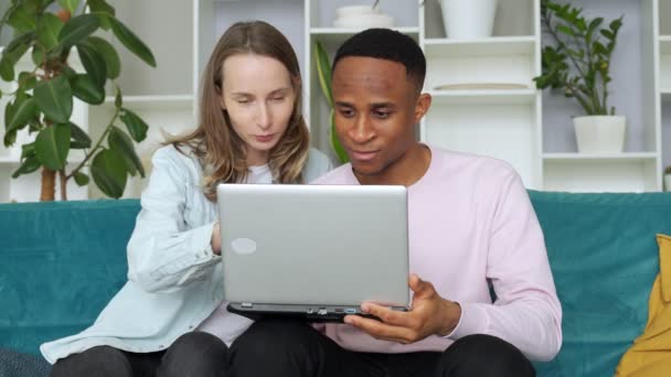 Χαρούμενη διαφυλετική ζευγάρι web surfing στο φορητό υπολογιστή μαζί, κάθεται στον καναπέ στο σπίτι — Αρχείο Βίντεο