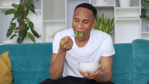 Hombre negro comiendo ensalada saludable sentado en el sofá. Concepto de alimentación saludable — Vídeos de Stock