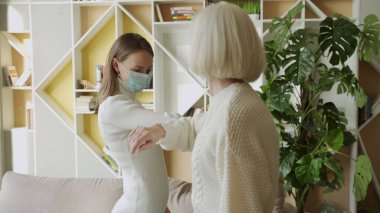 Ofiste dirseklerine dokunarak yüz maskesi takan yaşlı kadın ve genç kadın birbirlerini selamlıyorlar.