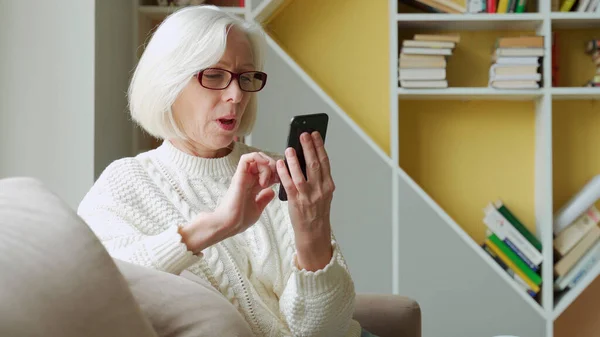 Mujer mayor está sentada en el sofá, sosteniendo un teléfono inteligente, sintiéndose muy feliz después de recibir una notificación por SMS sobre las noticias. Una anciana recibió un premio mientras sostenía un teléfono inteligente — Foto de Stock