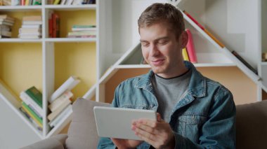 Tablet kullanan gülümseyen adam modern ofisteki koltukta dinleniyor. Evde çalışan genç iş adamları kavramı.