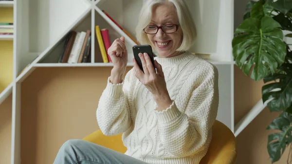 Mujer mayor está sentada en el sofá, sosteniendo un teléfono inteligente, sintiéndose muy feliz después de recibir una notificación por SMS sobre las noticias. Una anciana recibió un premio mientras sostenía un teléfono inteligente — Foto de Stock