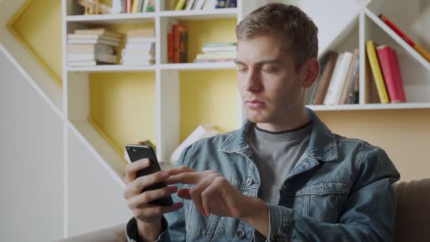 Il giovane sorridente tiene il telefono cellulare mentre è seduto sul divano a casa e festeggia — Video Stock