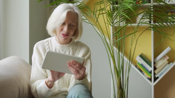 Mujer anciana usando una tableta de computadora, mirando a la pantalla, anciana anciana con canas comprando en línea usando aplicaciones, sentada en el sofá — Vídeo de stock