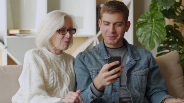 Gülümseyen orta yaşlı anne, yetişkin oğluyla birlikte akıllı telefon kullanıyor, aile hafta sonu, yaşlı anne cep telefonuyla kablosuz internete bakıyor.