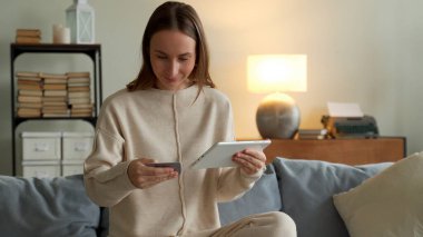 Oturma odasında online alışveriş yapmak için tablet kullanan gülümseyen bir kadın.