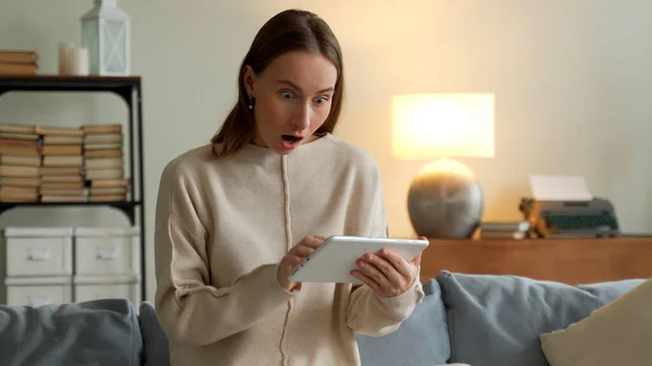 Woman at home on sofa, using tablet won playing online. Emotional young woman with tablet celebrating victory on sofa at home