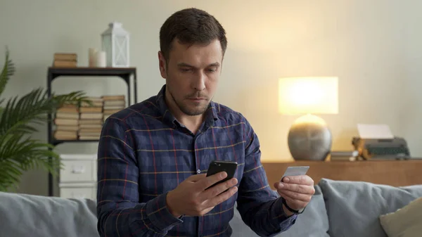 Красивый мужчина сидит дома на диване в гостиной со смартфоном и кредитной картой. Онлайн-оплата в интернет-магазине — стоковое фото