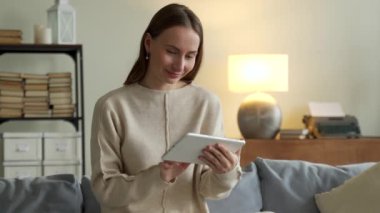 İnternette sörf yapan bir kadın. Çekici genç bir kadın dijital tablet kullanıyor ve evdeki kanepede otururken gülümsüyor.